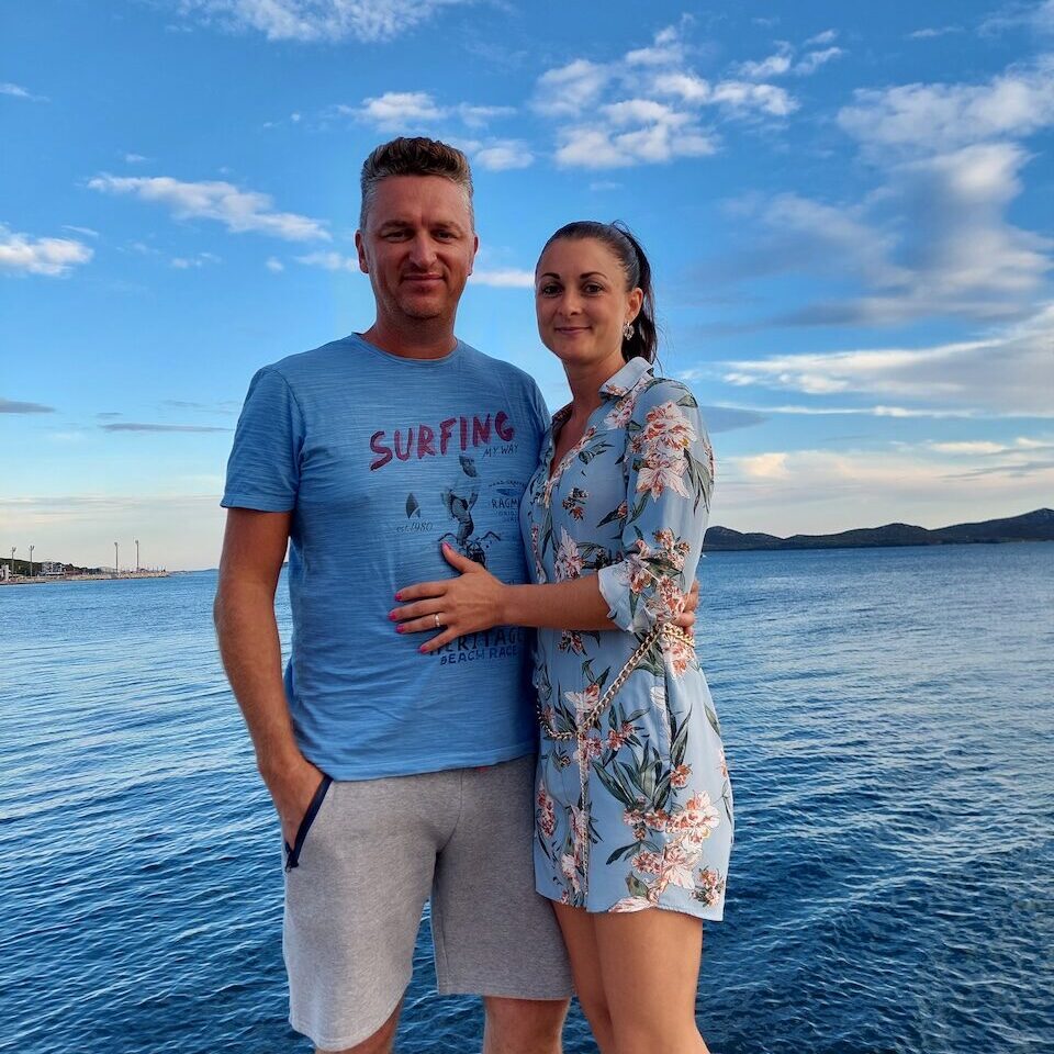 A smiling couple Petar on the left and Pascale on the right stands on a pier with blue sky and water in the background.