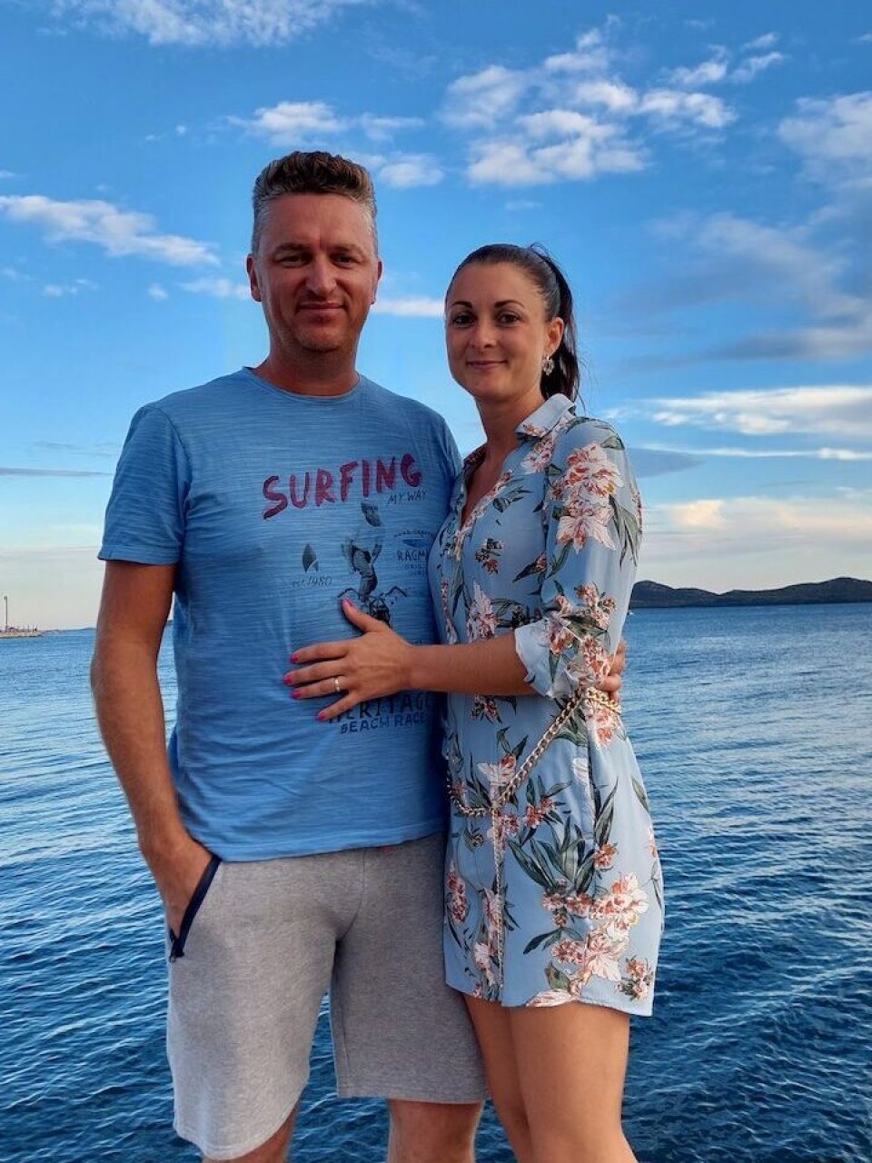 A smiling couple Petar on the left and Pascale on the right stands on a pier with blue sky and water in the background.